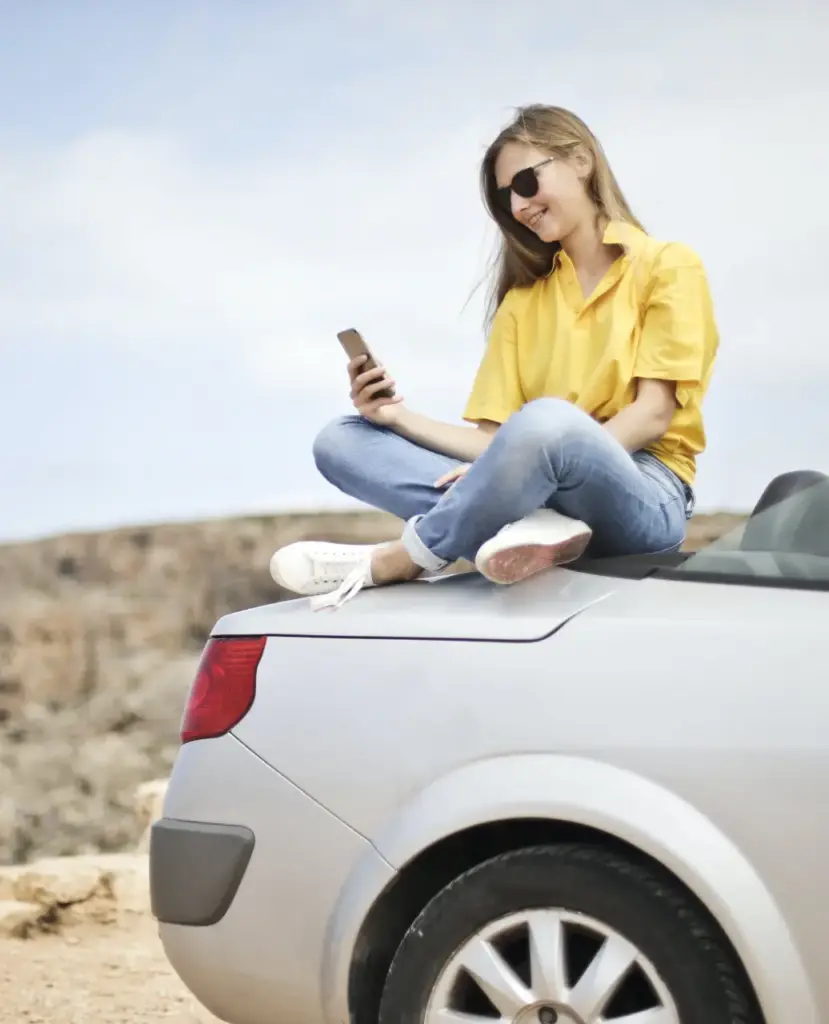woman sits on car browsing her phone, has a good car insurance